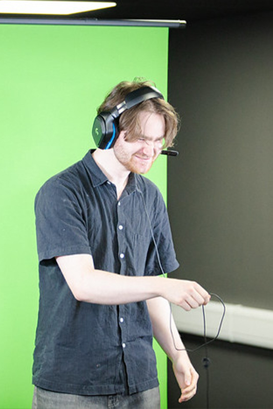 Students studying Esports. A student is stood in front of a green screen wearing a gaming headset and is posing.