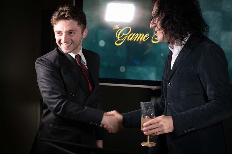 An image of two men in suits shaking hands, the left of which is lit well and is facing the camera and smiling having received an award.